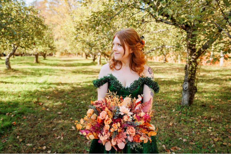 Bride in an emerald green gown holding a faux rust, burgundy and orange bridal bouquet in an orchard