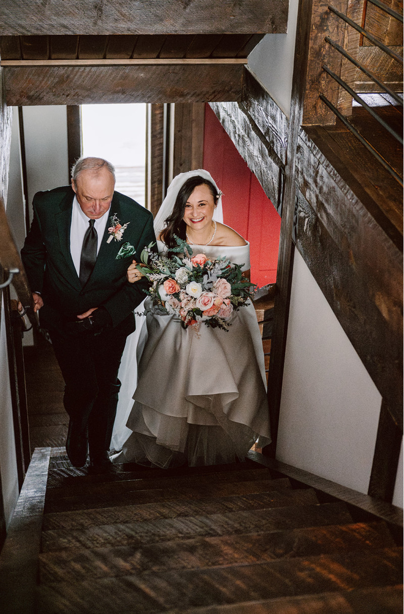 Bride walking down staircase with peach, blush pink and greenery wedding bouquet and father