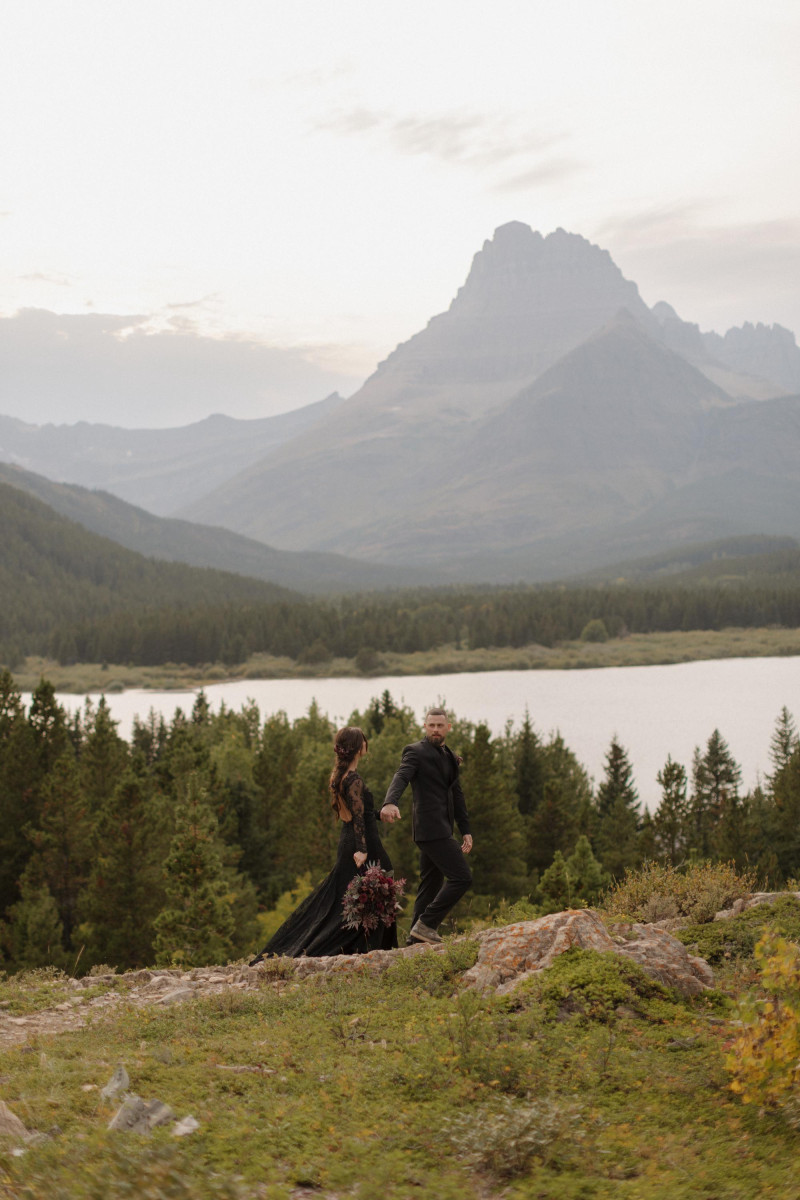 purple, burgundy and gold wedding couple in black attire walking in mountain landscape with lake and forest backdrop