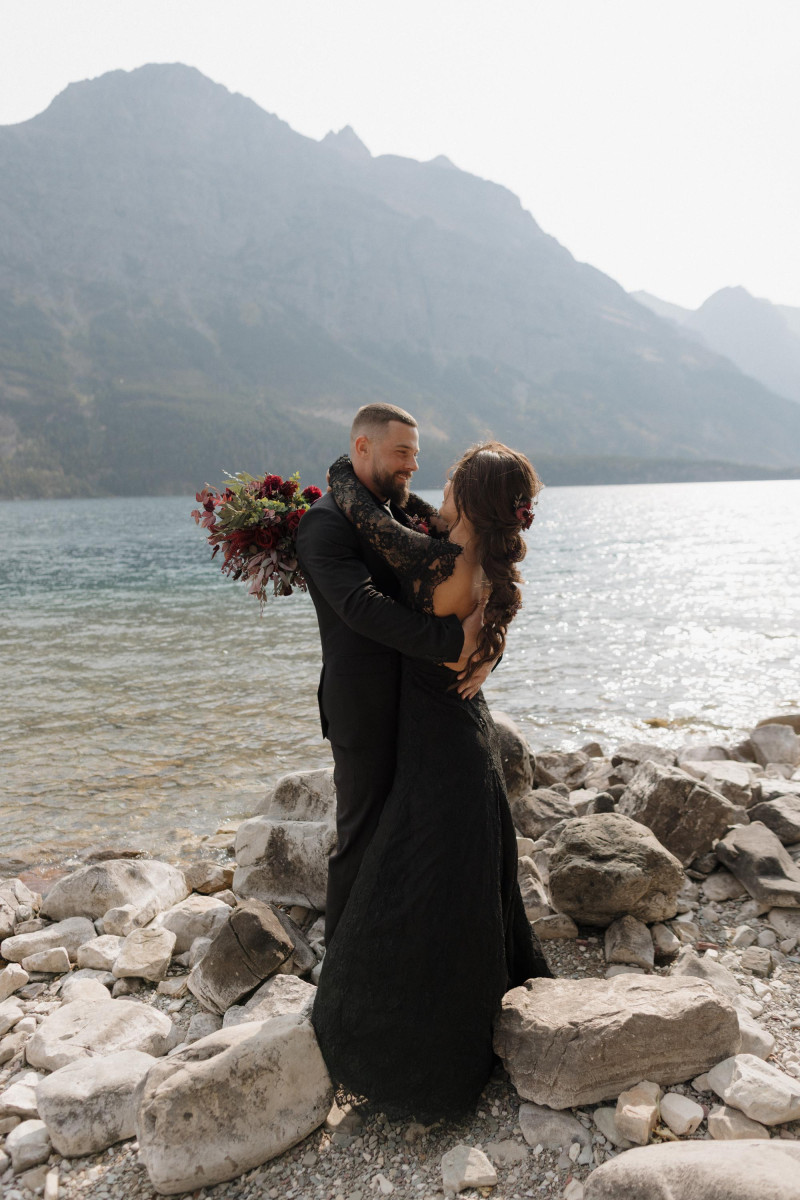 bride and groom in black embracing by the lake with purple, burgundy and gold wedding bouquet